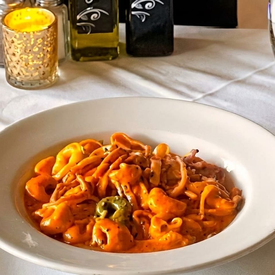 A white bowl of tortellini in a creamy orange sauce on a white tablecloth with a candle and water glass.