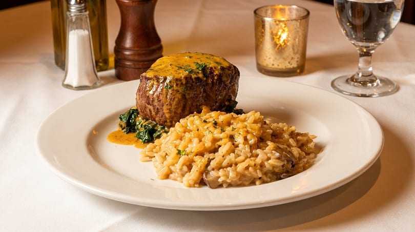 A filet mignon with sauce, spinach, and risotto on a white plate, with a candle and water glass.