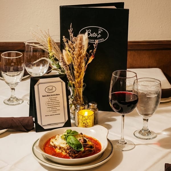 A Beto's Italian Bistro table set with lasagna, red wine, water, and menus.