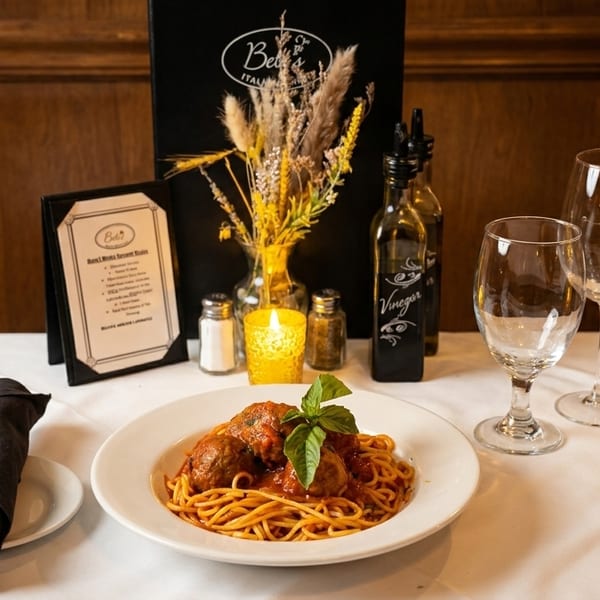 Spaghetti and meatballs with basil on a white plate, set on a table with wine glasses and menu.